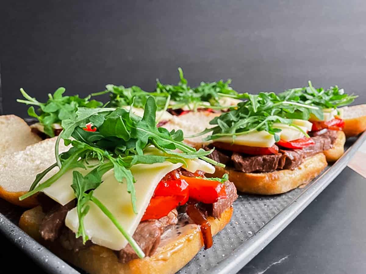 Assembly of steak sandwich piled with caramelized onion and pepper, arugula, and gruyere cheese.