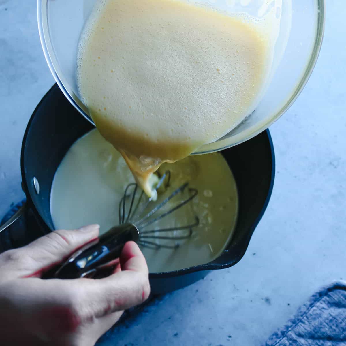 Process shot of tempering the eggs in which the warm milk mixture is poured into the yolks and then poured back into the pan before finishing cooking.